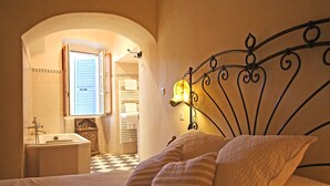 Un dormitorio con cama, mesita de noche, espejo y baño visible a través de un arco.