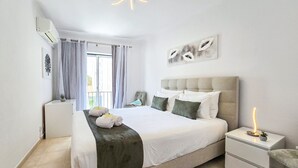 A bedroom with a bed, bedside tables, a chair, a window with curtains, a ceiling light, and a wall-mounted artwork. - Apartment Praia Rocha Justar Cozy (Portimão)