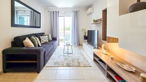 A modern living room with a sofa, TV, and a small table with a lamp. - Apartment Praia Rocha Justar Cozy (Portimão)