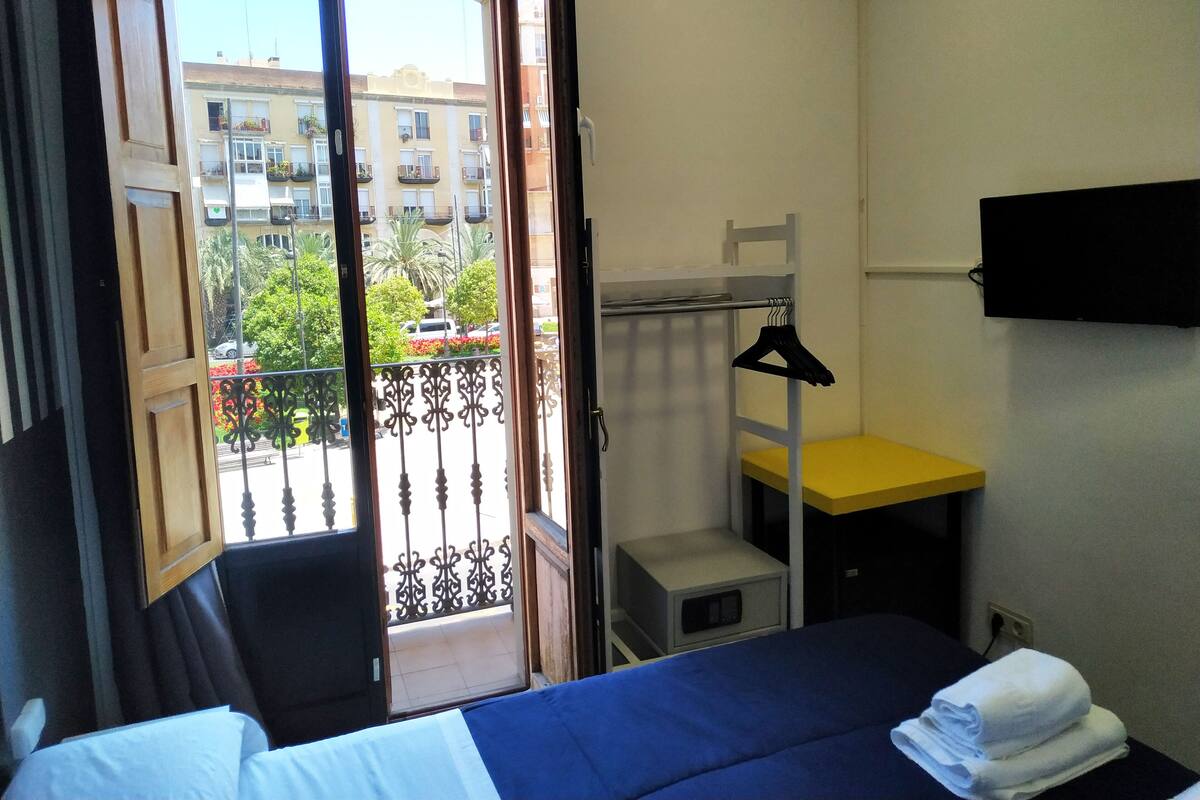 Een hotelkamer met een bed, een klein bureau, een televisie en een balkon met uitzicht op gebouwen en bomen.