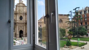 Vista a través de una ventana de un edificio histórico con arquitectura ornamentada, un patio con bancos y una estatua.