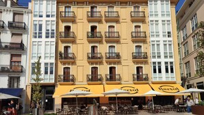 Un edificio de varios pisos con fachada amarilla, varios balcones y zonas de estar exteriores con sombrillas.