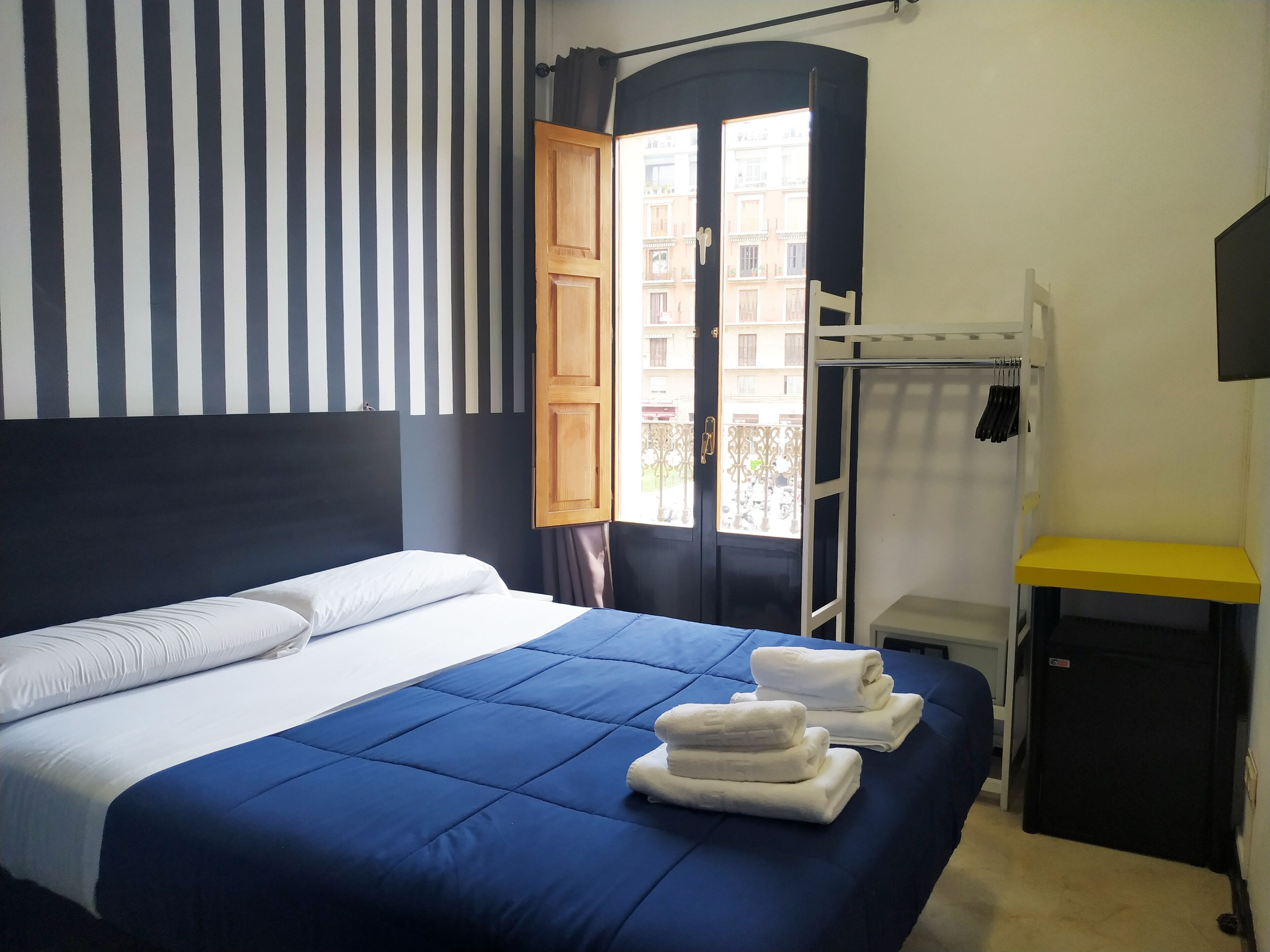 Habitación de hotel con cama, armario de madera, una mesita y vistas a edificios desde la puerta.