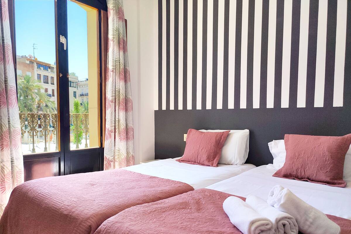 Een hotelkamer met twee bedden, een groot raam met gordijnen, een gestreept behang en uitzicht op gebouwen en bomen.