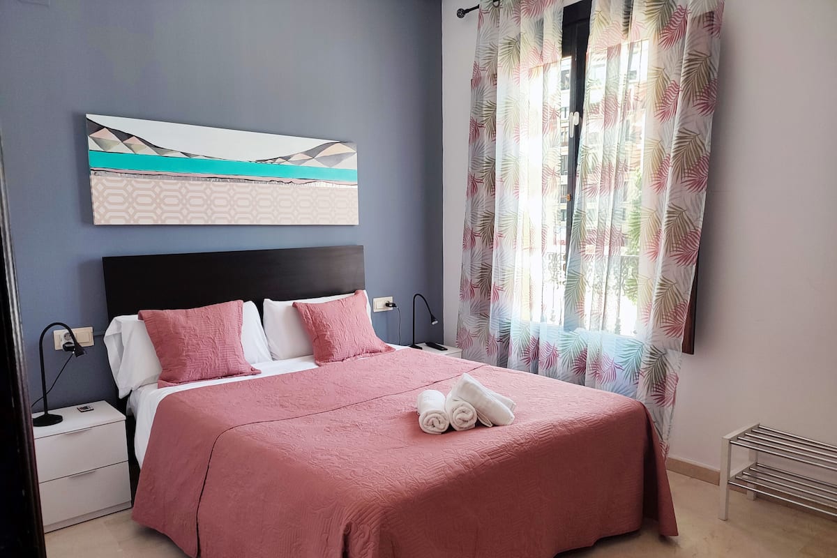 Een net opgemaakt bed met roze beddengoed, een hoofdbord en een kunstwerk aan de muur erboven.
