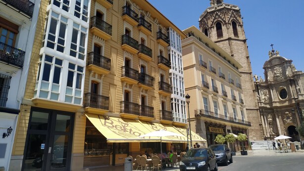 Un edificio con varios balcones, un café con terraza y una iglesia histórica al fondo.