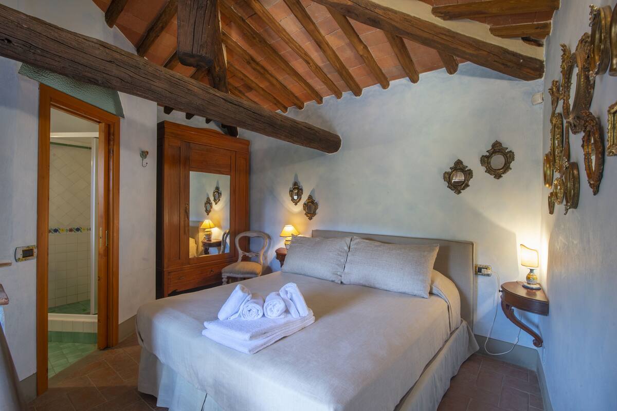 A bedroom with a bed, a wooden ceiling, a bathroom with a shower, and a wall with decorative mirrors.