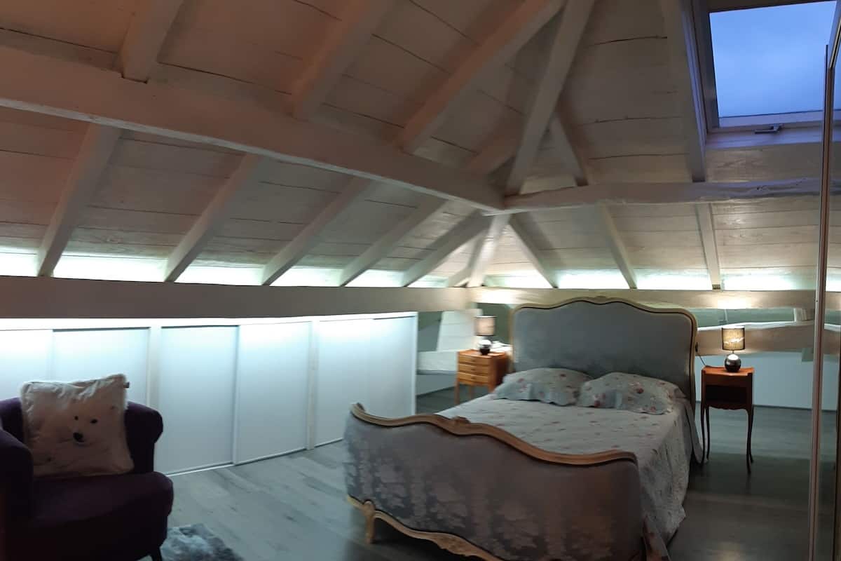 Quarto com teto abobadado, cama com roupa de cama floral, mesa de cabeceira com abajur e cadeira com almofada.