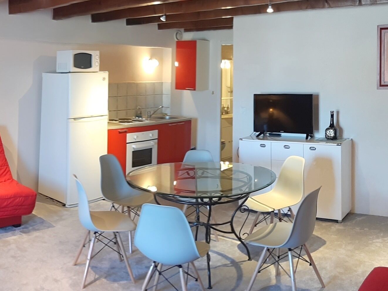 A compact kitchen with a microwave, oven, and red cabinets. A glass table with chairs is in the center, and a TV is on a cabinet to the right.