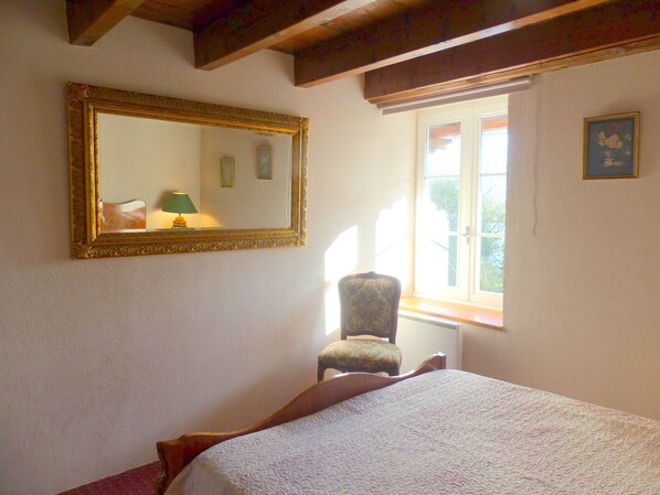 Un dormitorio con cama, una silla, un ventanal y un espejo en la pared.
