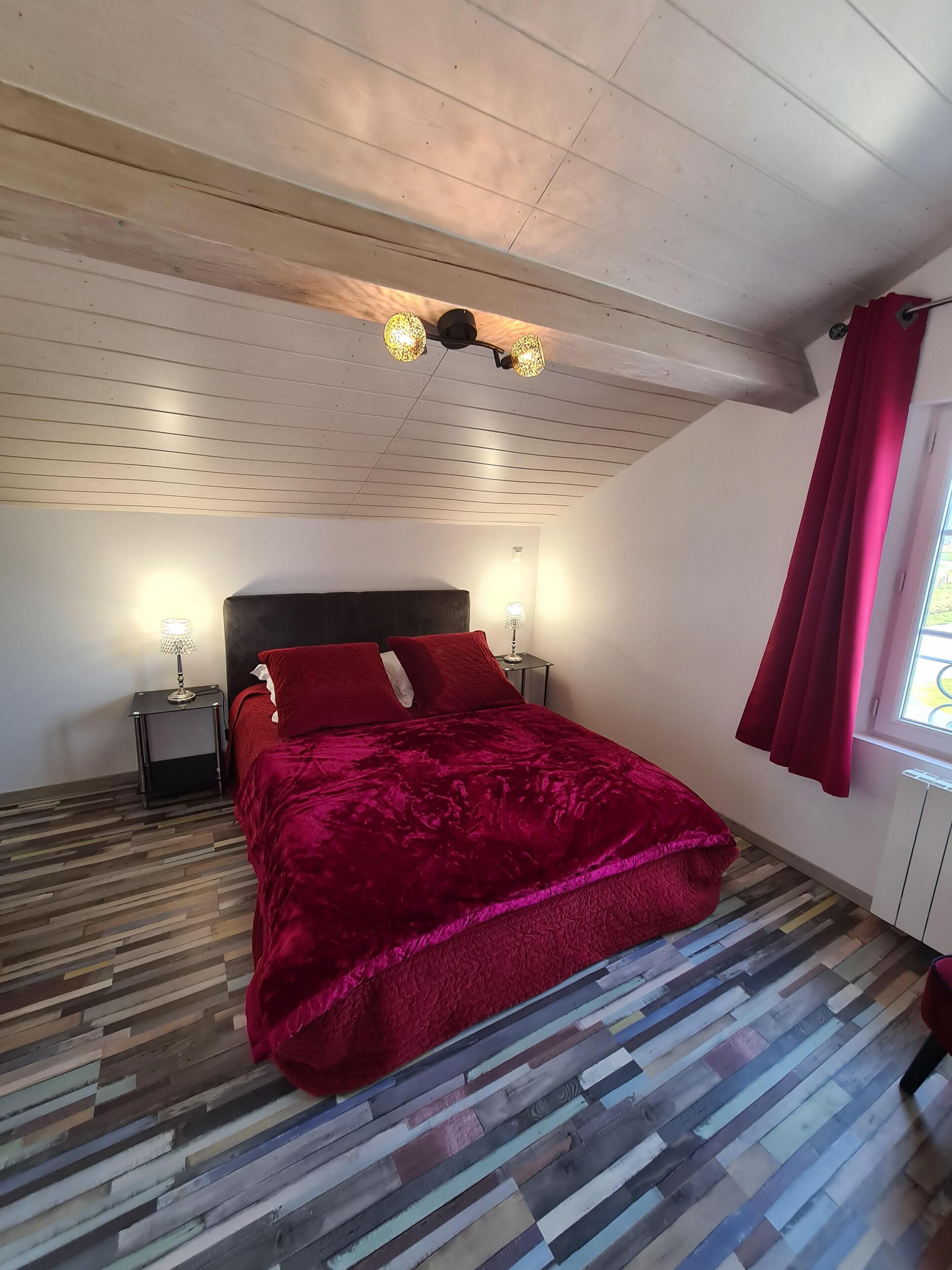 Un dormitorio con techo de madera, una cama con colcha roja, dos lámparas de noche y una ventana con cortinas rojas.