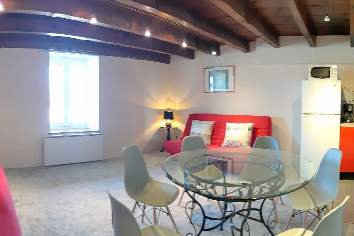 Uma sala de jantar com mesa e cadeiras de vidro, um sofá vermelho com almofadas, um abajur e uma geladeira branca.