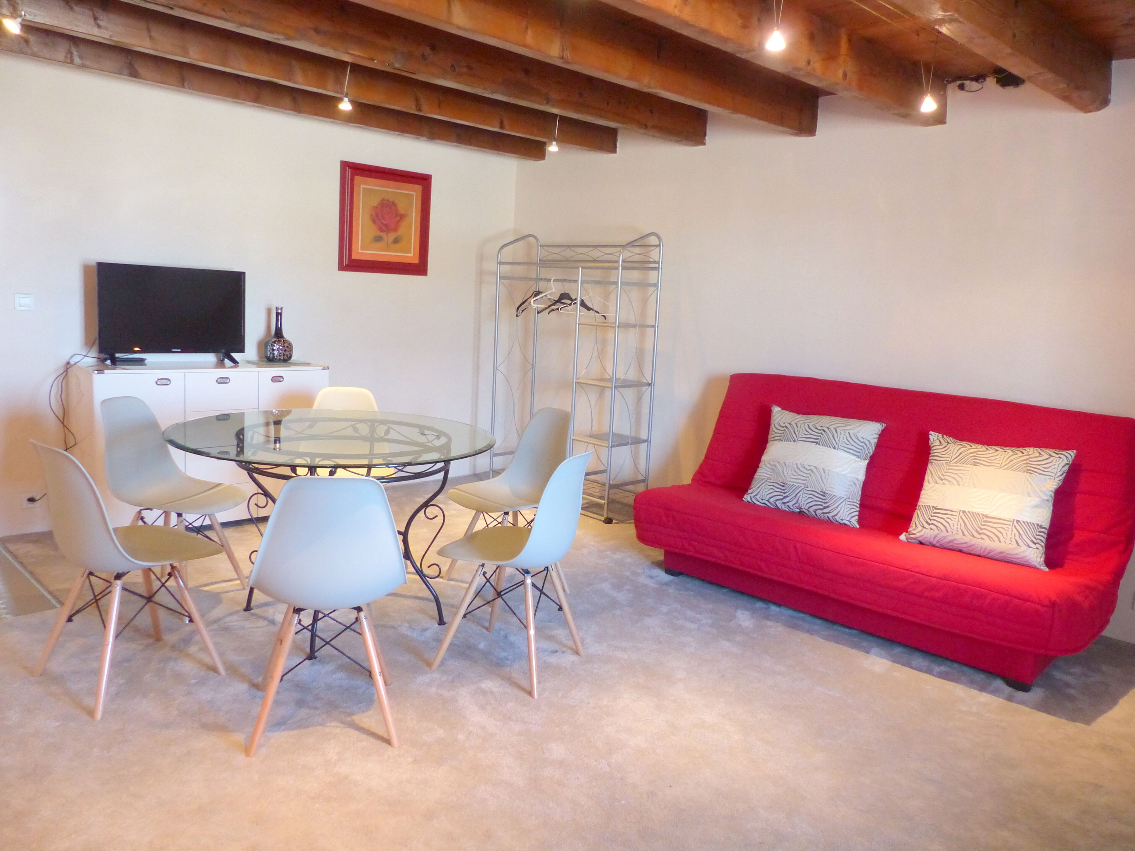 Une pièce comprenant une table ronde en verre entourée de quatre chaises, un canapé rouge orné de coussins à motifs, un téléviseur posé sur un meuble blanc et une étagère en métal.