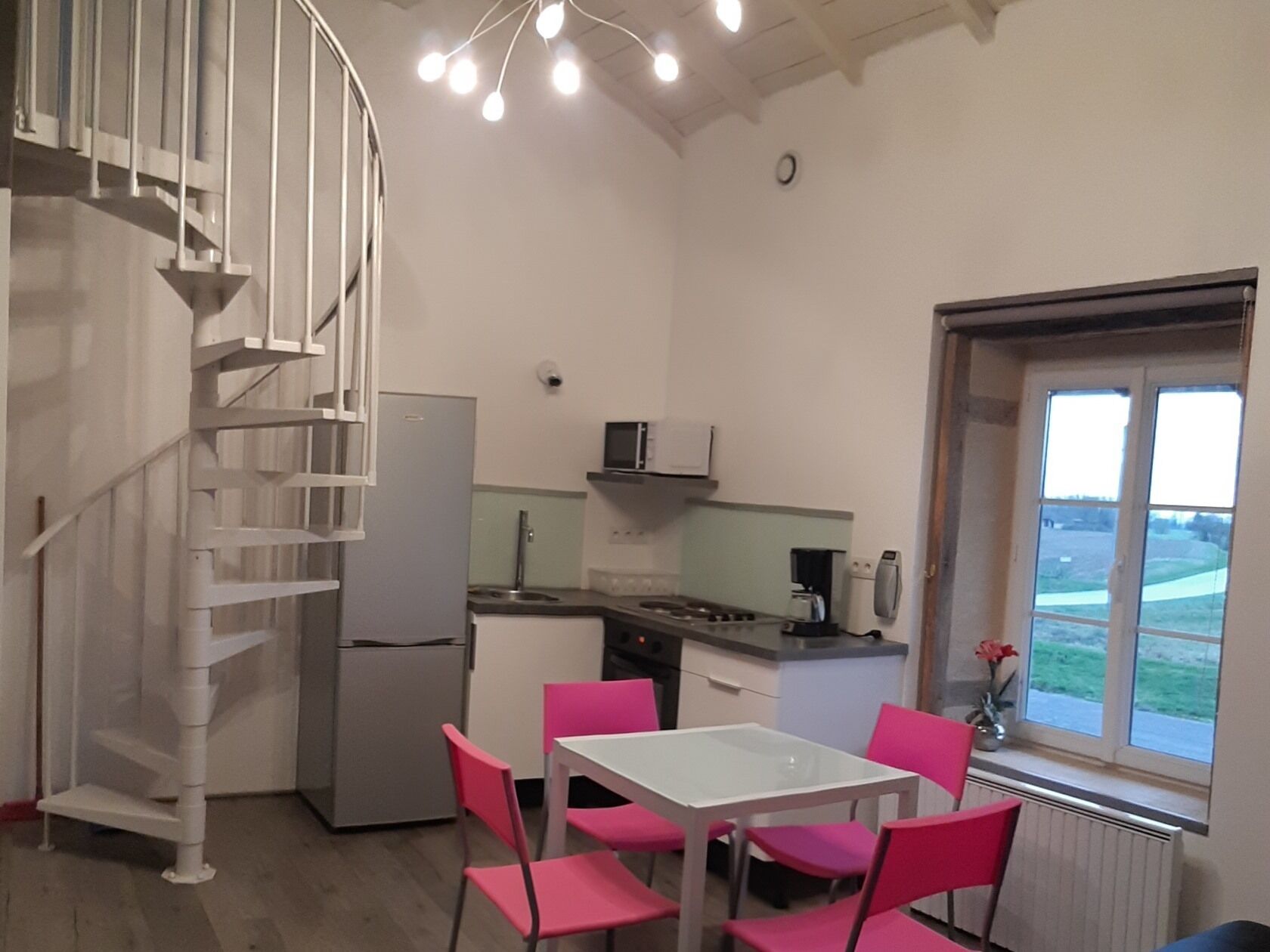 A compact kitchen with a white table and chairs, a spiral staircase, and a window with a view.