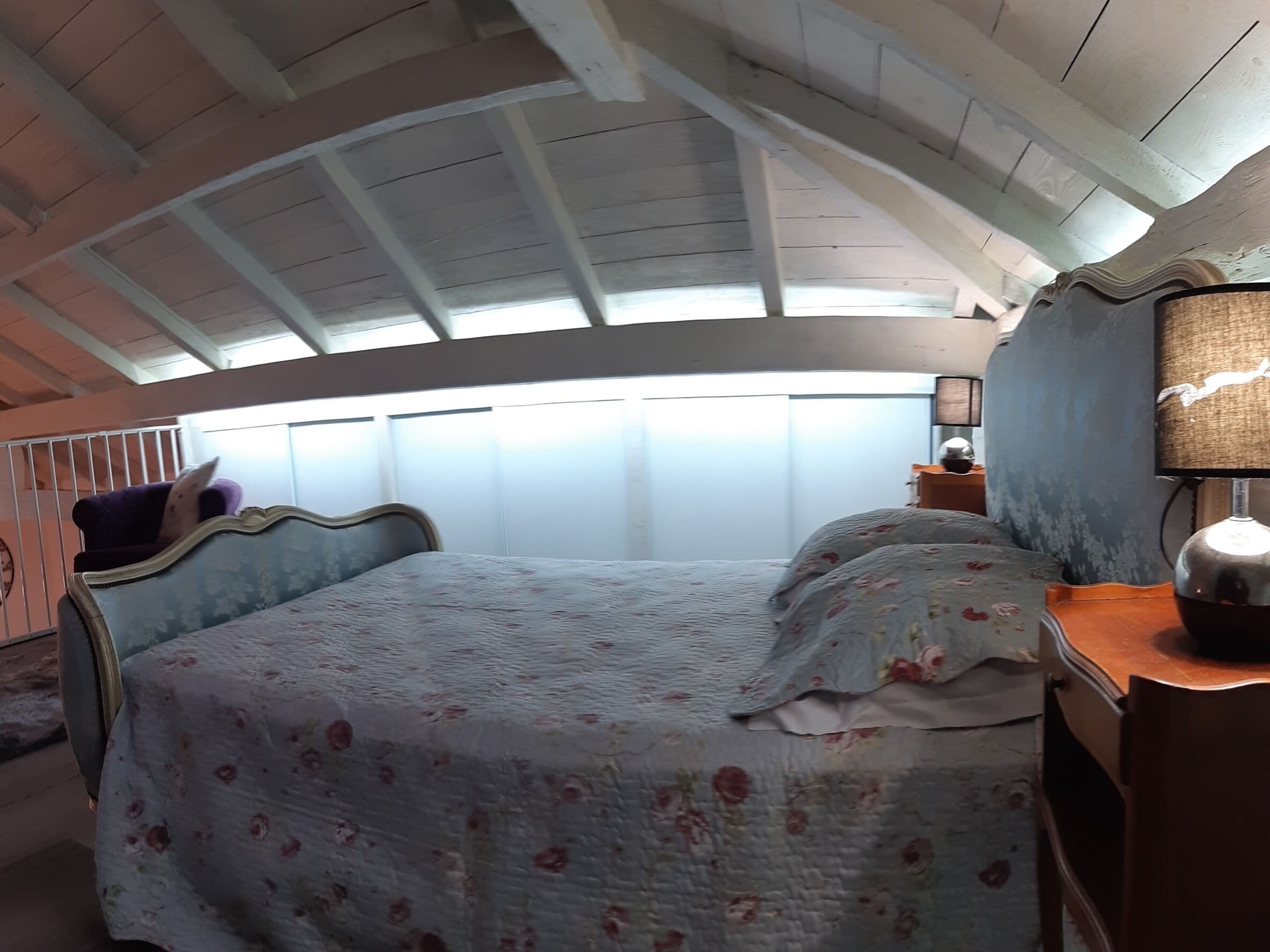 A bedroom with a curved ceiling, a bed with floral bedding, a nightstand with a lamp, and a window with blinds.