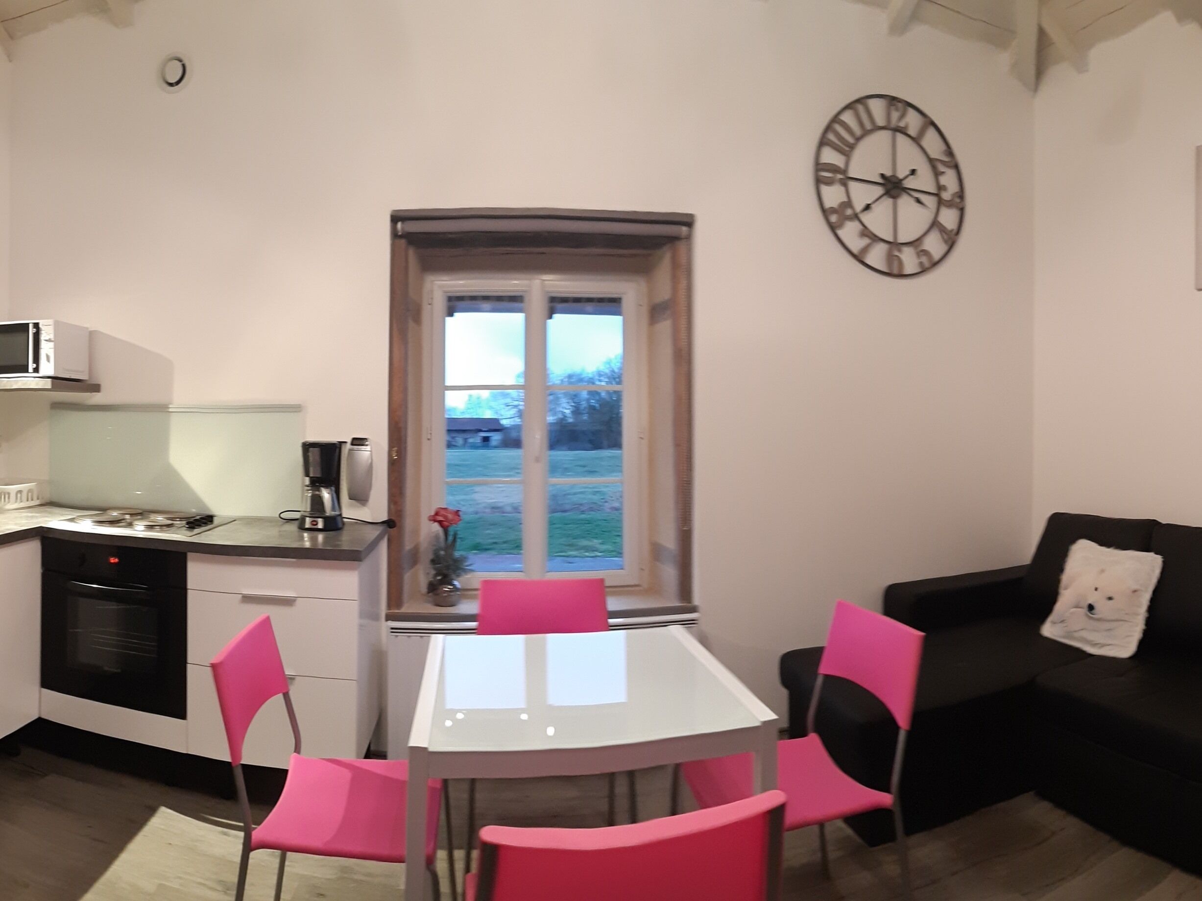 A compact living space with a kitchenette, a dining table with chairs, a black sofa, and a large window with a view of the outdoors.