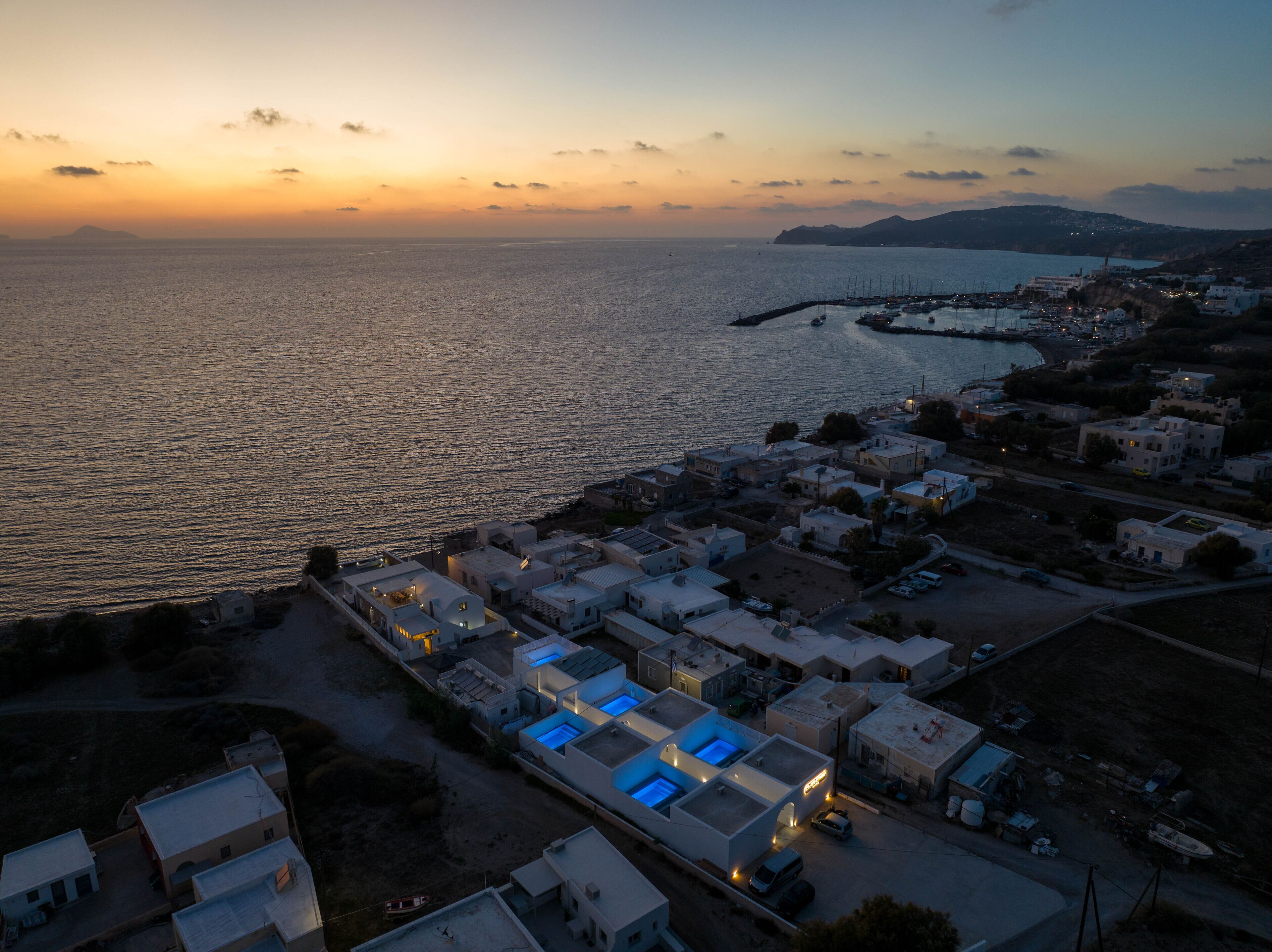 An aerial view of a coastal town at sunset with a marina, buildings, and a clear view of the sea.