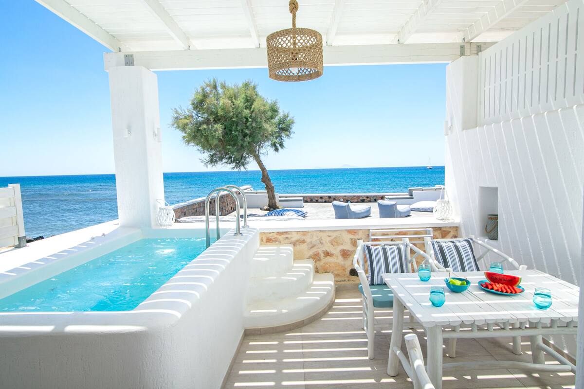 A pool area with a view of the sea, a white table with blue cushions, and a hanging light fixture.