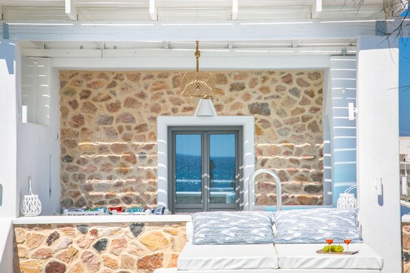 A balcony with a stone wall, a door, and a bed with a view of the sea. - Aegean Melody Suites (Santorini)