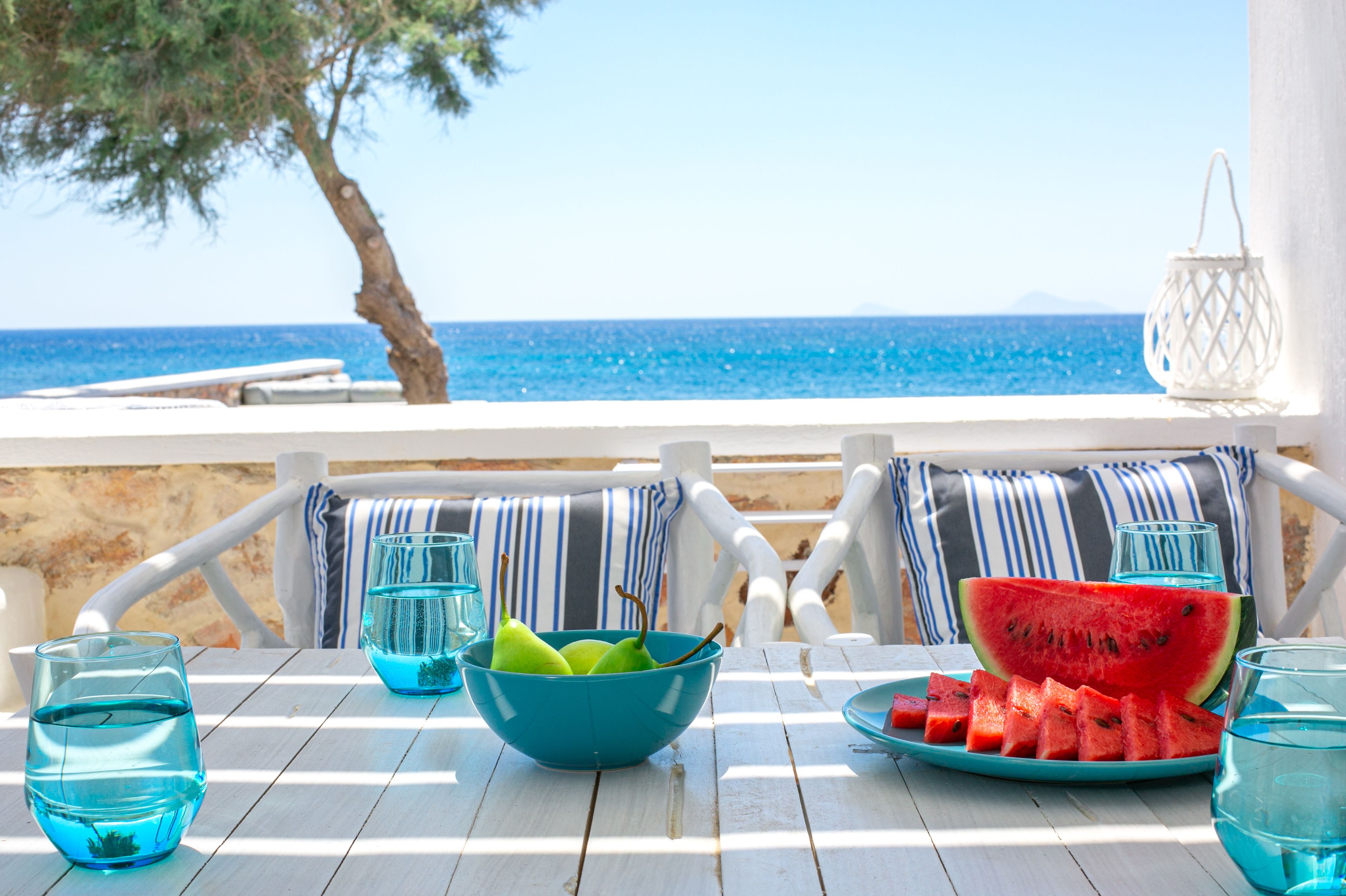 Una mesa de madera blanca con un tazón azul, rebanadas de sandía y vasos, frente a una vista al mar.