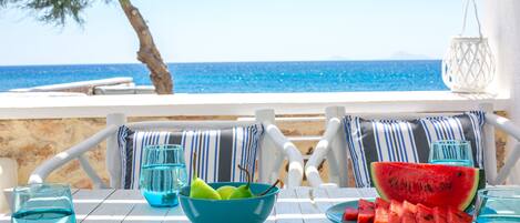 Una mesa de madera blanca con un tazón azul, rebanadas de sandía y vasos, frente a una vista al mar.