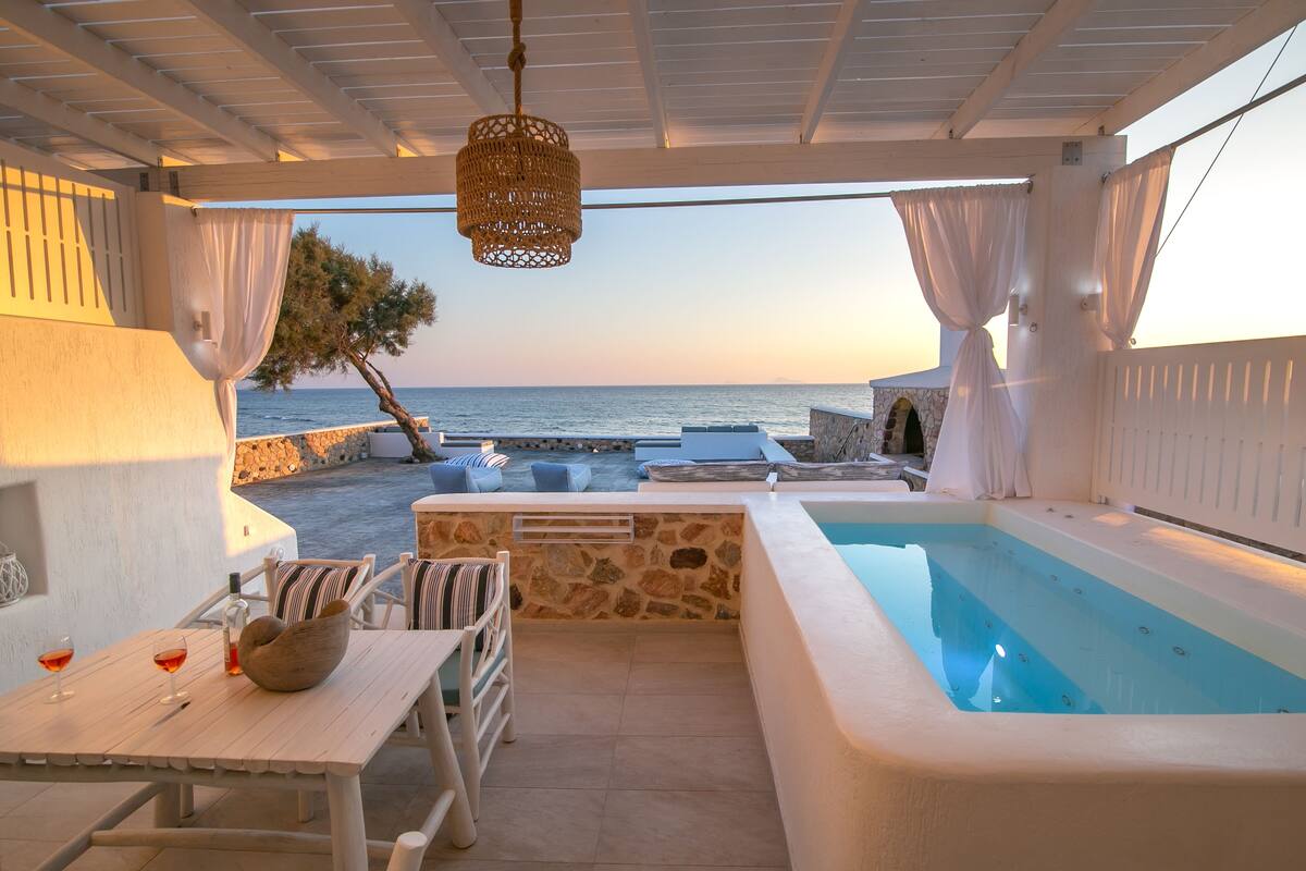 An outdoor pool area with a stone wall, a table with chairs, and a view of the sea at sunset.