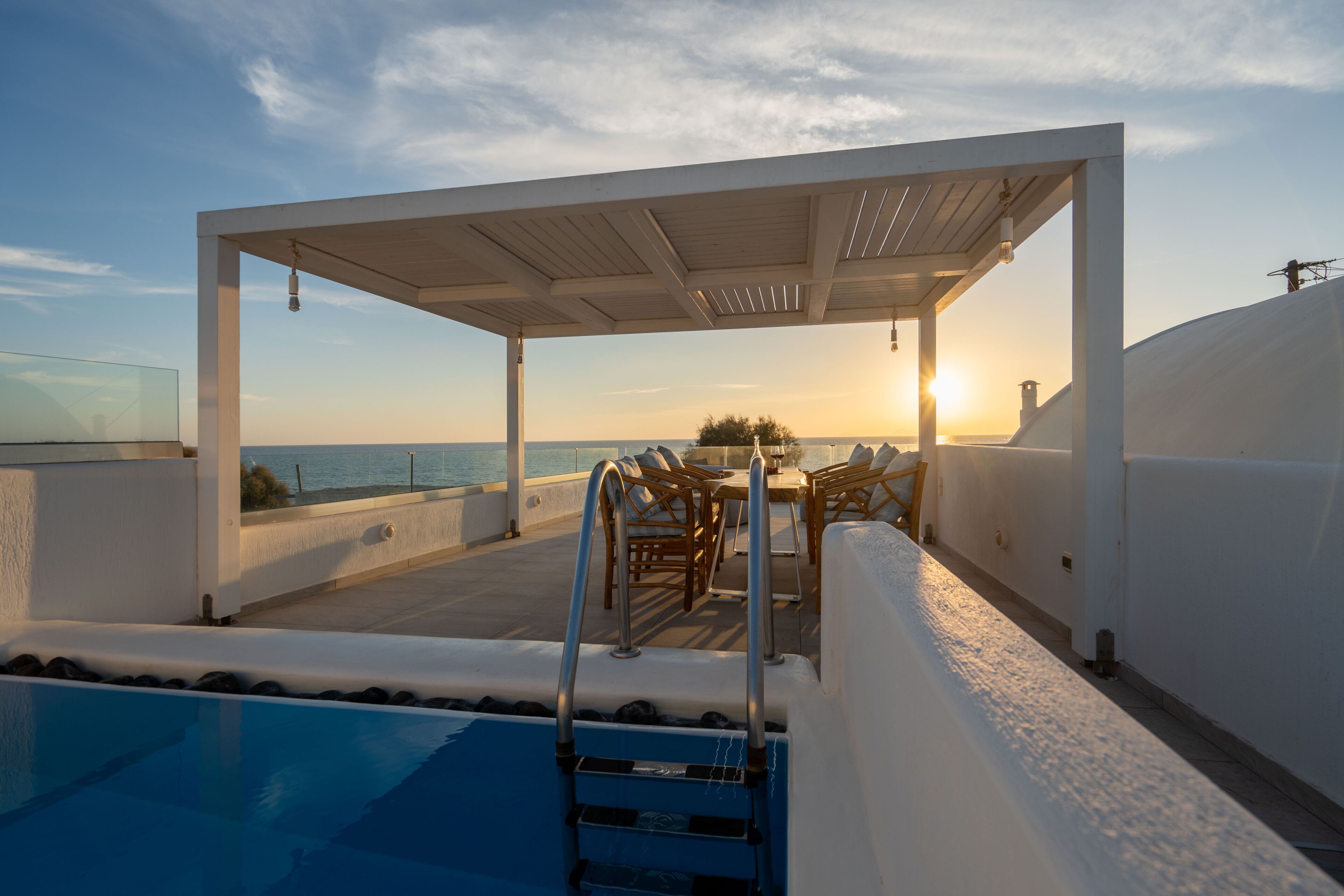 A rooftop pool area with a pergola, lounge chairs, and a view of the sunset over the ocean.
