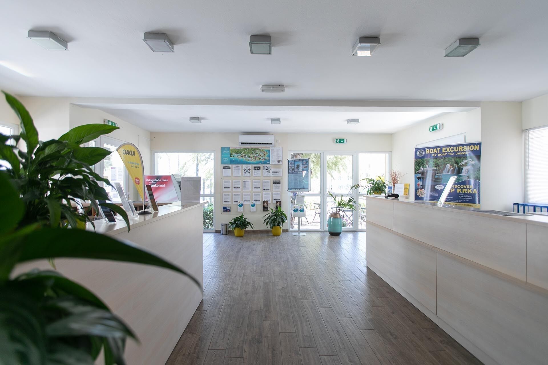 A reception area with a wooden floor, potted plants, a reception desk, informational posters, and a clear view to the outside.