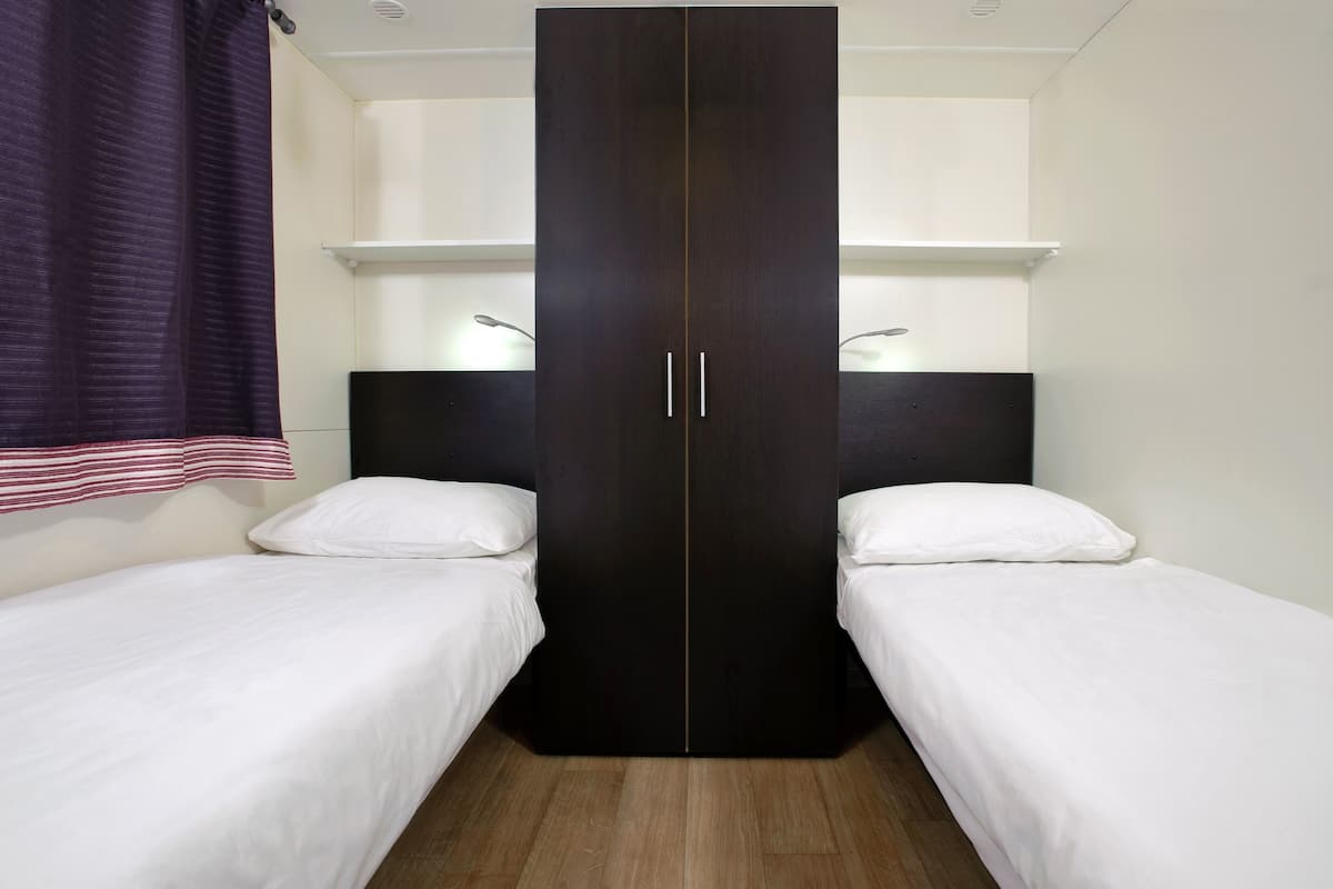 Ein Zimmer mit zwei Einzelbetten, einem dunklen Kleiderschrank und einem Fenster mit Vorhängen.