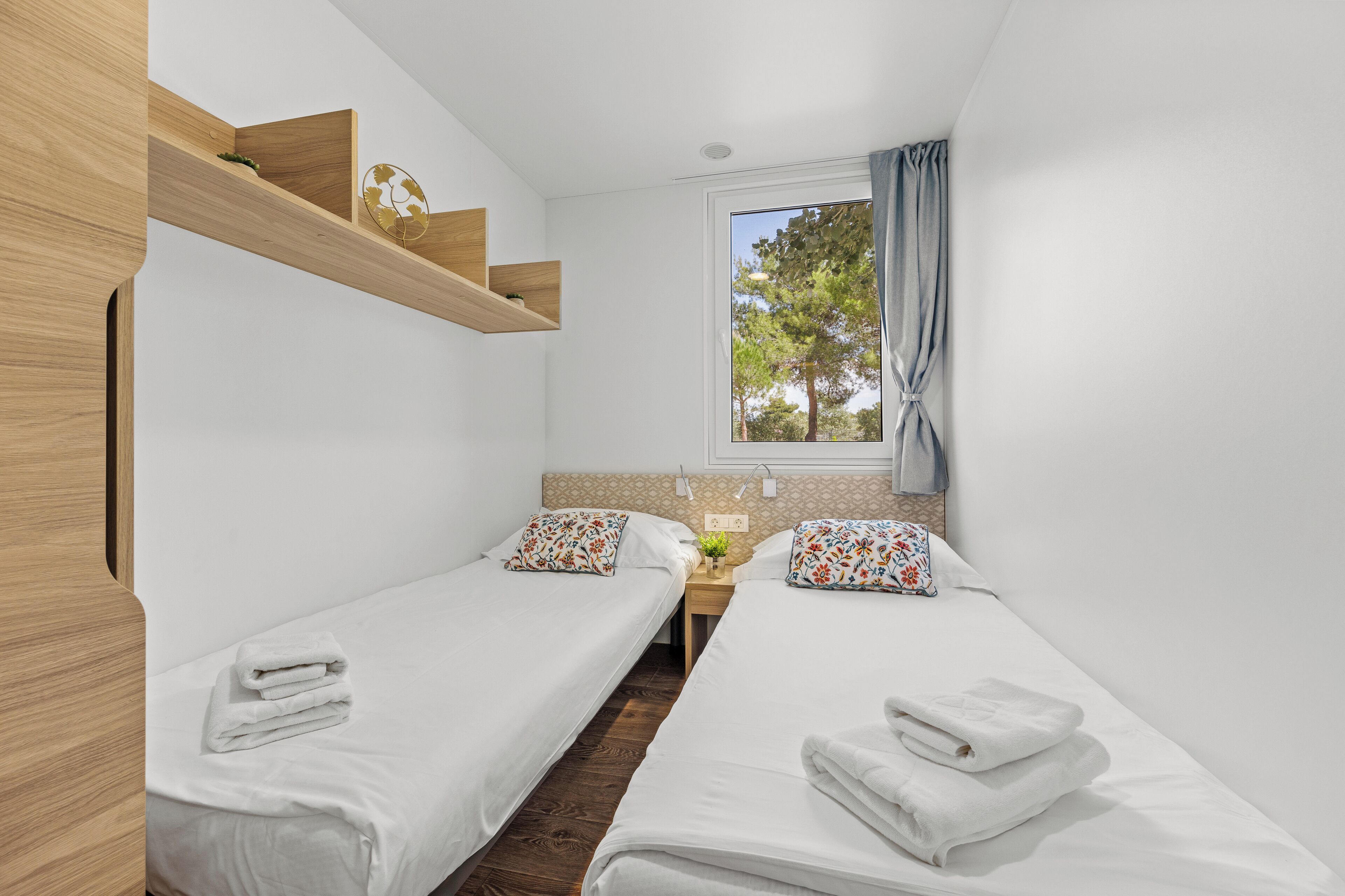 Une petite chambre moderne avec deux lits simples, une étagère en bois, une fenêtre donnant sur des arbres et une petite table avec un vase.