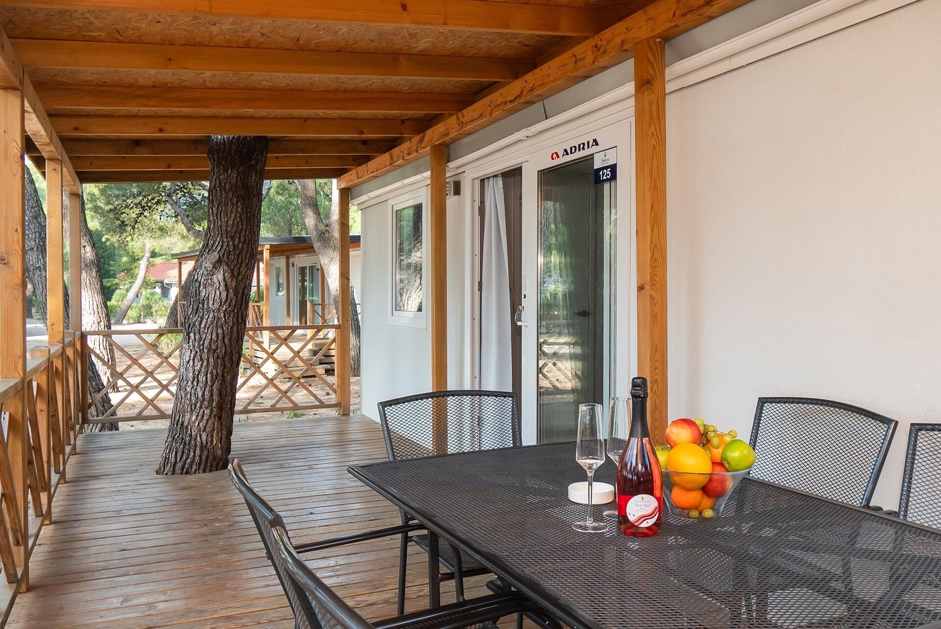 A covered outdoor seating area with a table set for two, featuring a bottle of red wine and a bowl of fruit.
