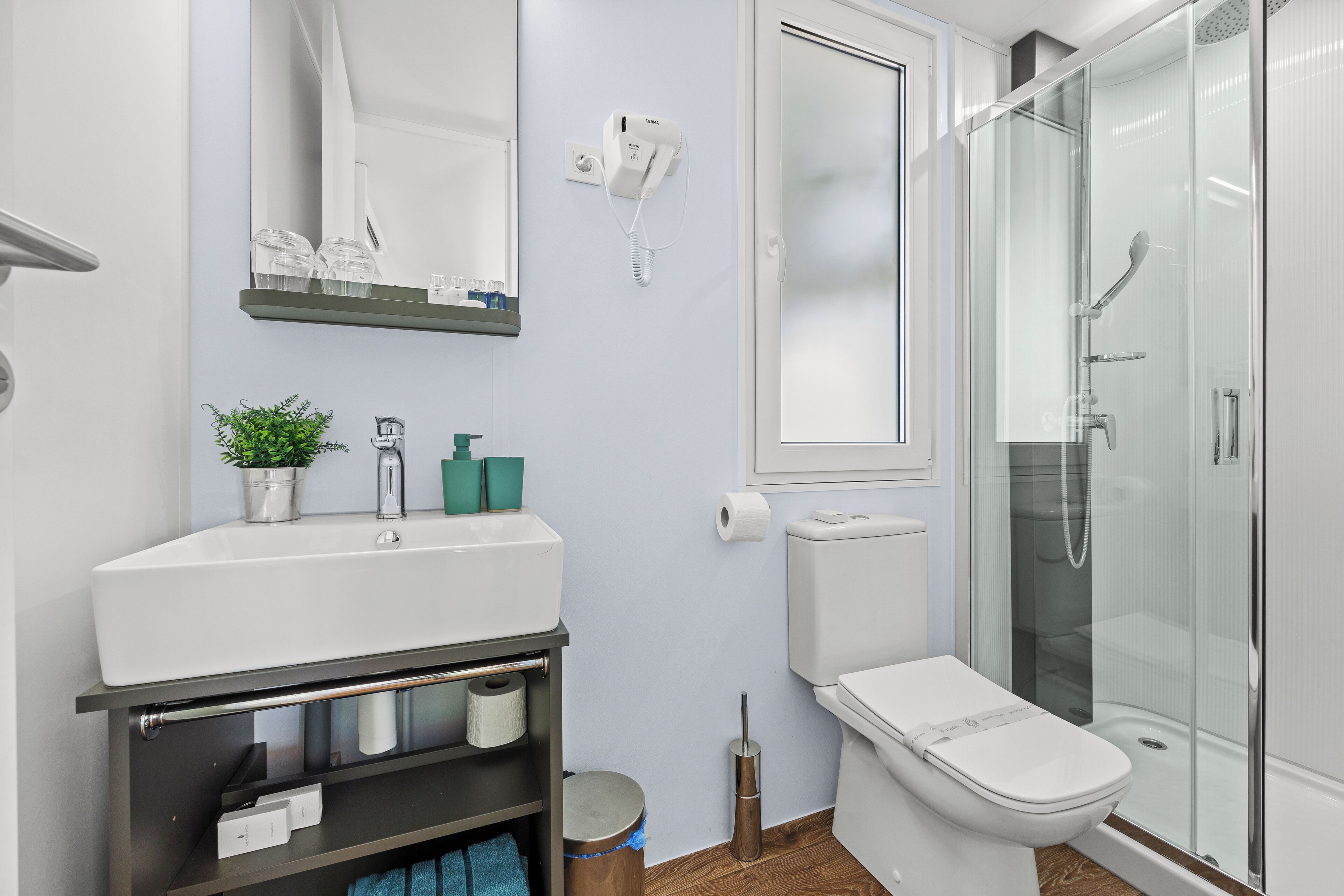 Un baño moderno con lavamanos, espejo y retrete blancos, cabina de ducha de vidrio y un secador de pelo montado en la pared.