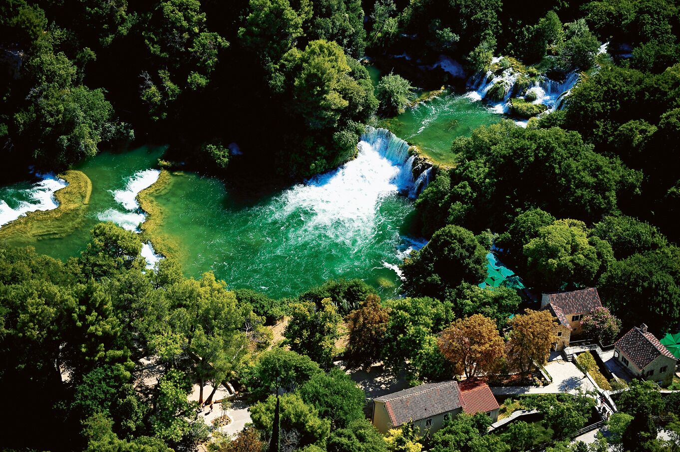 Luftaufnahme eines Flussufers mit einem Wasserfall, dichtem Grün und traditionellen Gebäuden mit roten Ziegeldächern.
