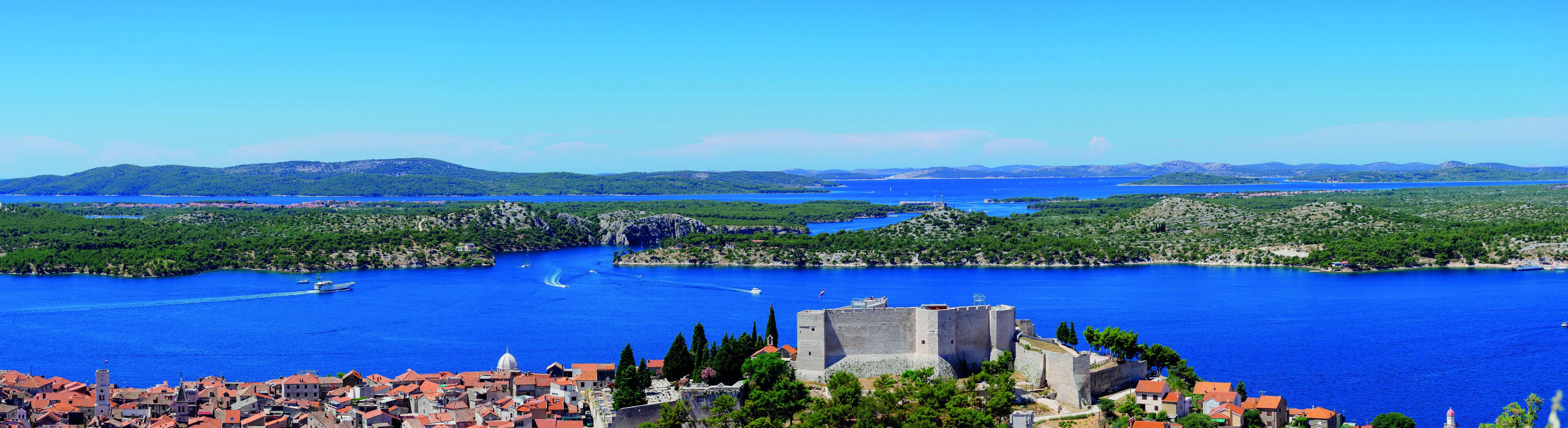 Un pueblo costero con un castillo histórico, aguas azul turquesa y colinas circundantes.