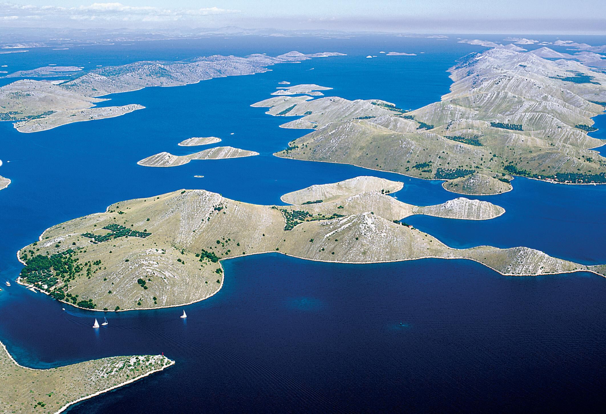 Luftaufnahme einer Küstenlandschaft mit zahlreichen Inseln und Buchten, darunter klares, blaues Wasser und spärliche Vegetation.