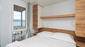 A bedroom with a wooden headboard, a bed with white linens, a window with a view of the sea, and a small table with a chair outside.