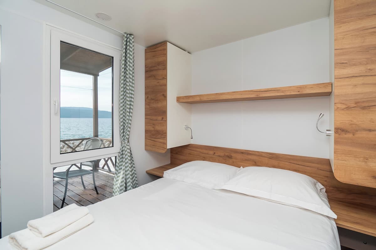 Ein Schlafzimmer mit hölzernem Kopfteil, einem Bett mit weißen Leinentüchern, einem Fenster mit Meerblick und einem kleinen Tisch mit Stuhl davor.