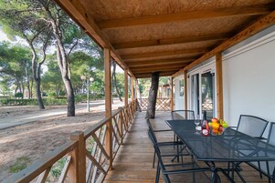 A covered outdoor seating area with a table and chairs, a wooden deck, and a view of trees and a building in the background.