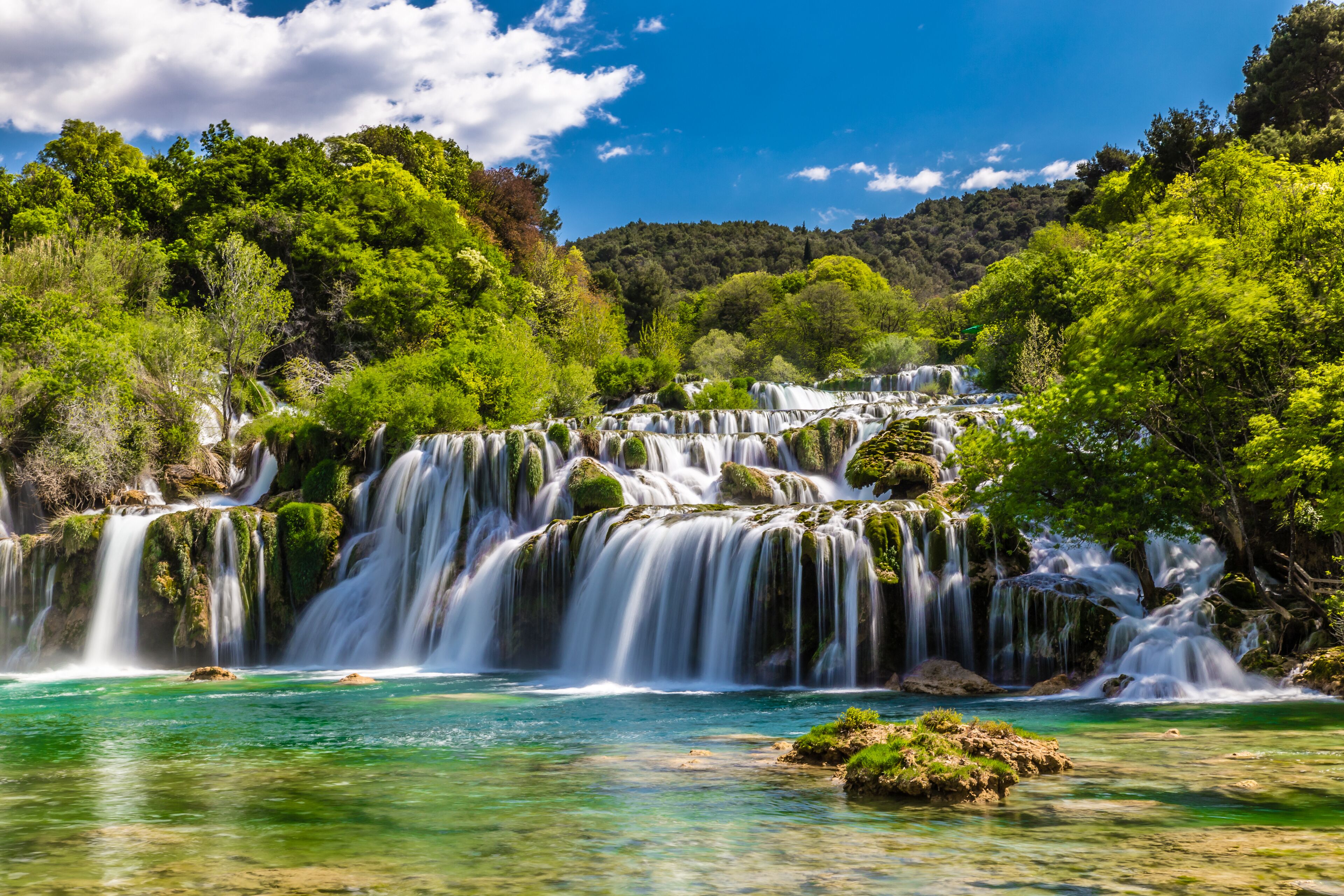 Una cascada se precipita sobre rocas hacia una piscina clara de color turquesa, rodeada de exuberante vegetación.