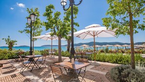 An outdoor patio with tables and chairs, surrounded by umbrellas and trees, overlooking a body of water and hills.