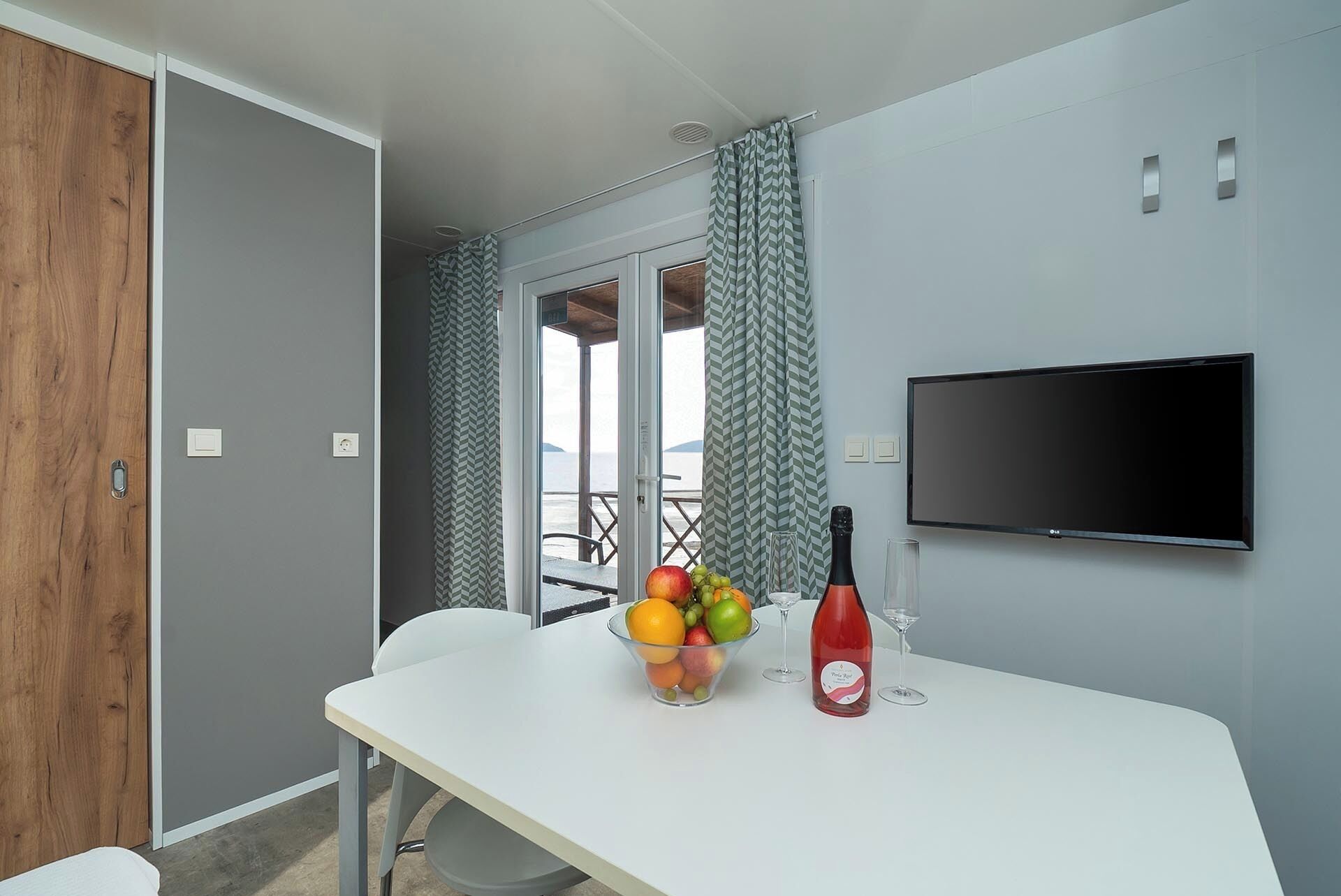 Une pièce moderne avec une table blanche, un panier de fruits, une bouteille de vin et un téléviseur fixé au mur.