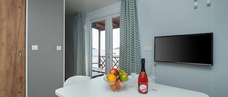 Une pièce moderne avec une table blanche, un panier de fruits, une bouteille de vin et un téléviseur fixé au mur.