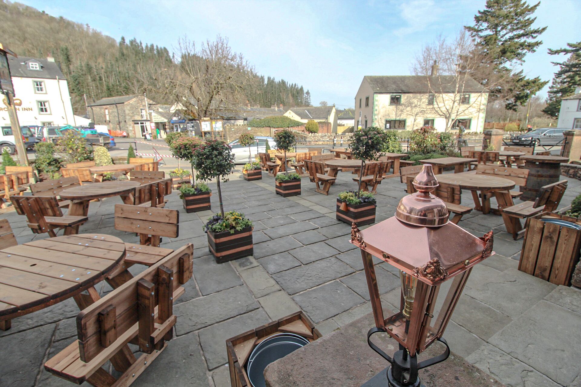 An outdoor seating area with wooden tables and benches, surrounded by potted plants and a decorative lantern.