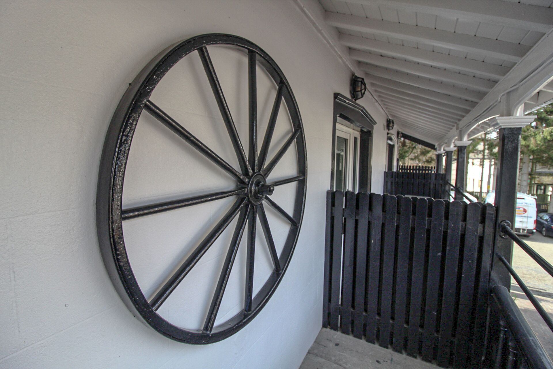 A large black wheel mounted on a white wall, a wooden fence, and a covered porch area.