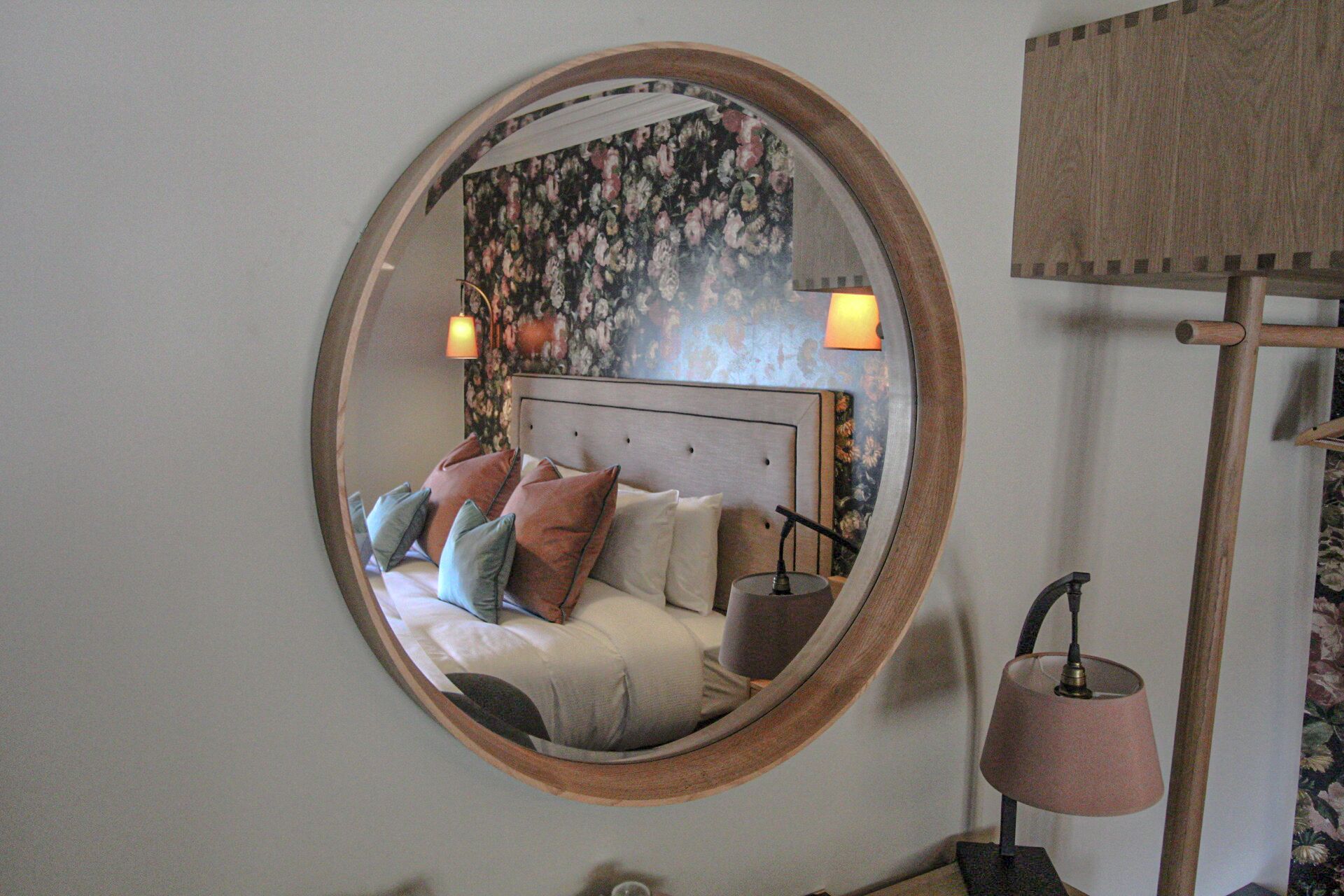 A bedroom with a round mirror, a bed with pillows, a nightstand, and a lamp.