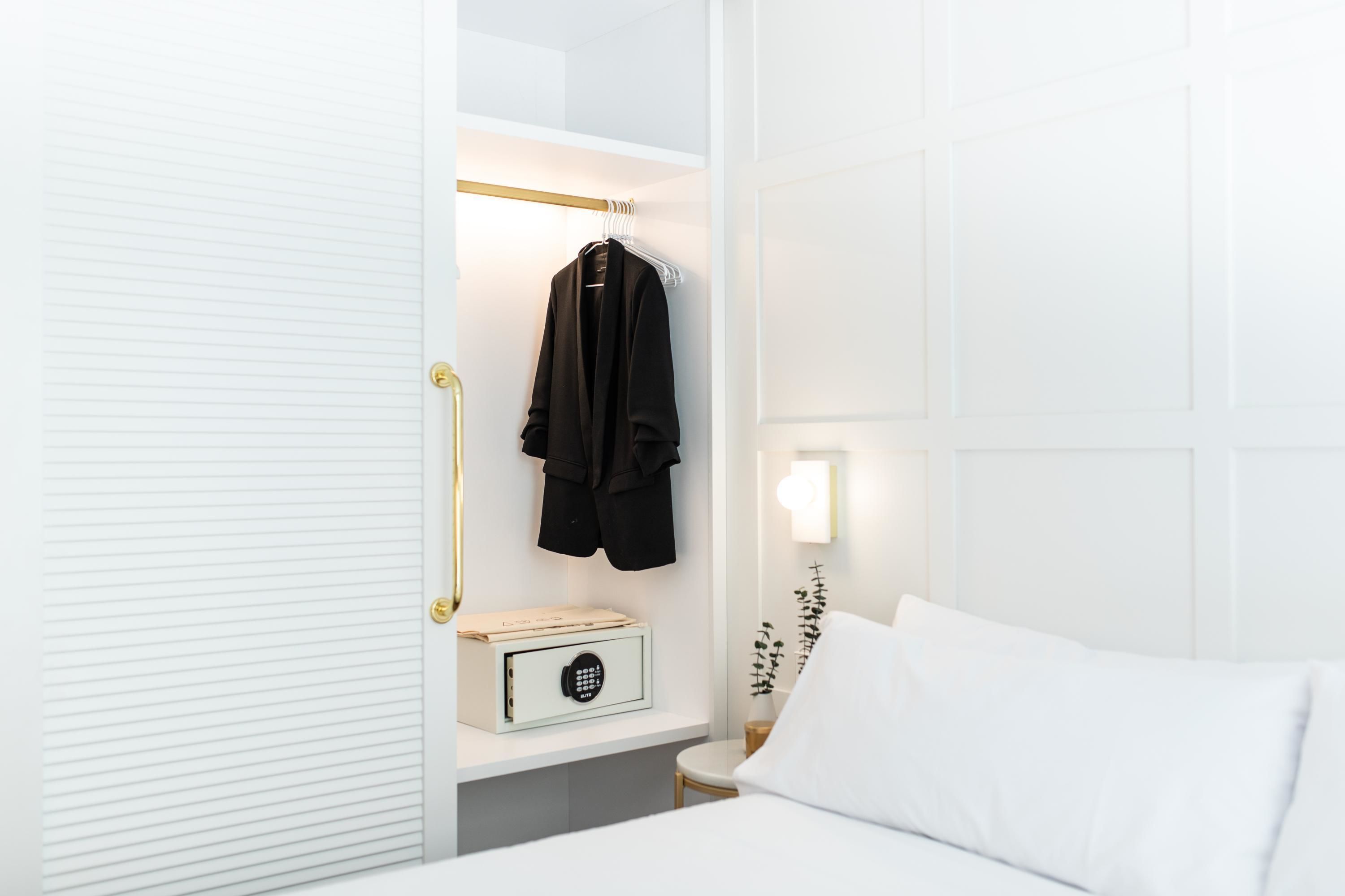 Et hotelværelse med en hvid seng, et natbord med en lampe, en garderobe med en frakke og et pengeskab.