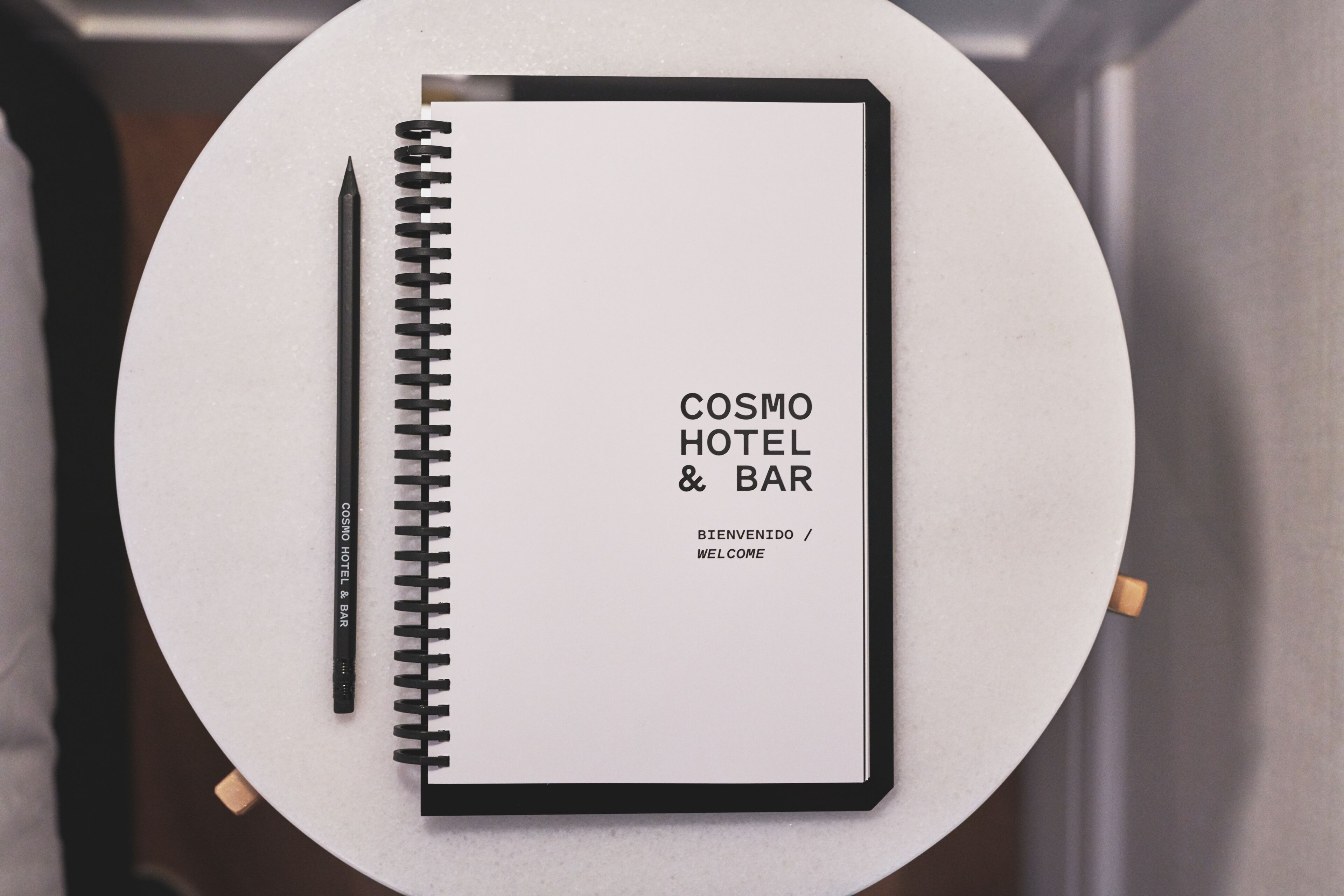 Un cuaderno con la etiqueta "COSMO HOTEL & BAR" y "BIENVENIDO / WELCOME" en la portada, sobre una mesa redonda.