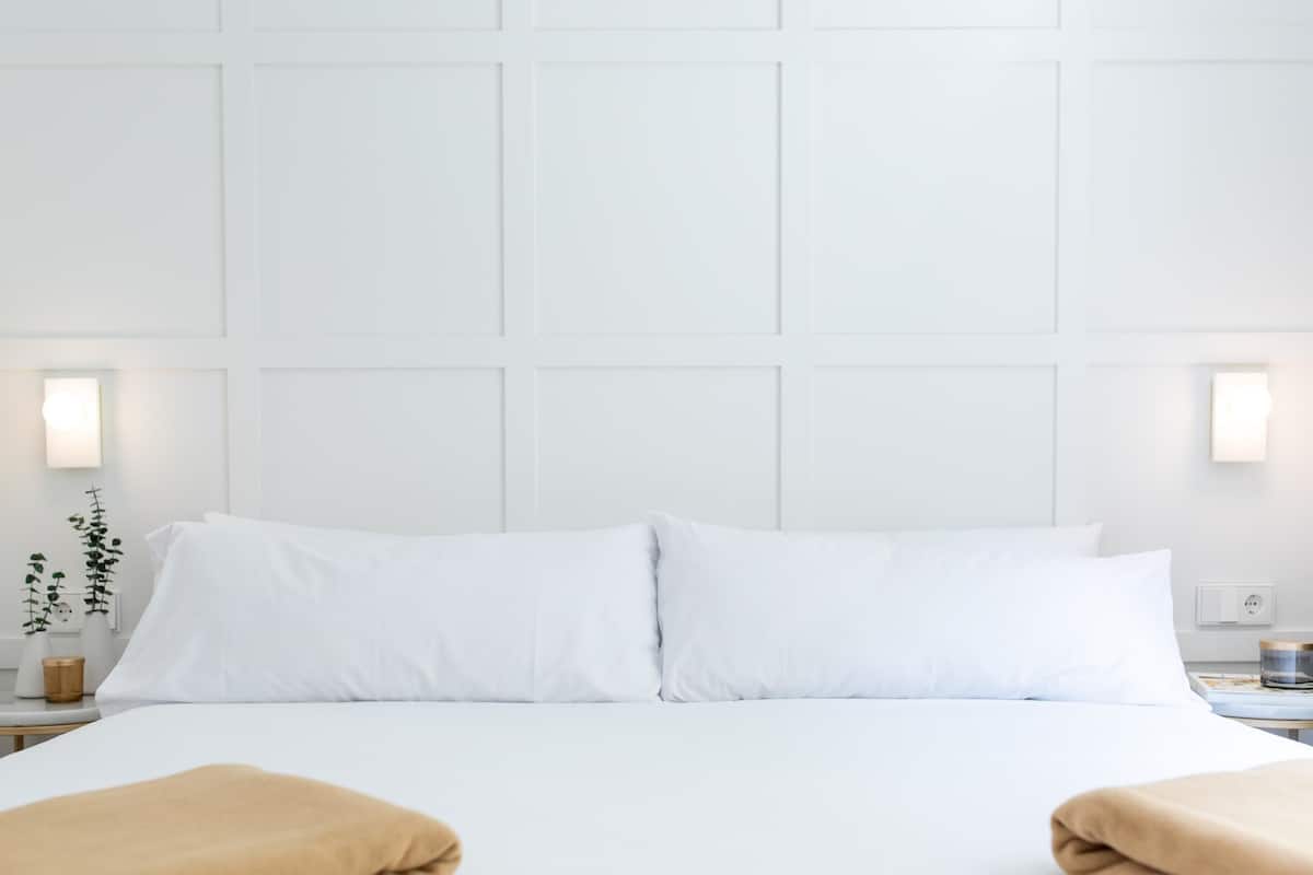 Habitación de hotel con una cama grande, ropa de cama blanca, dos lámparas de noche, una mesita de noche con una planta y una lámpara fijada en la pared.