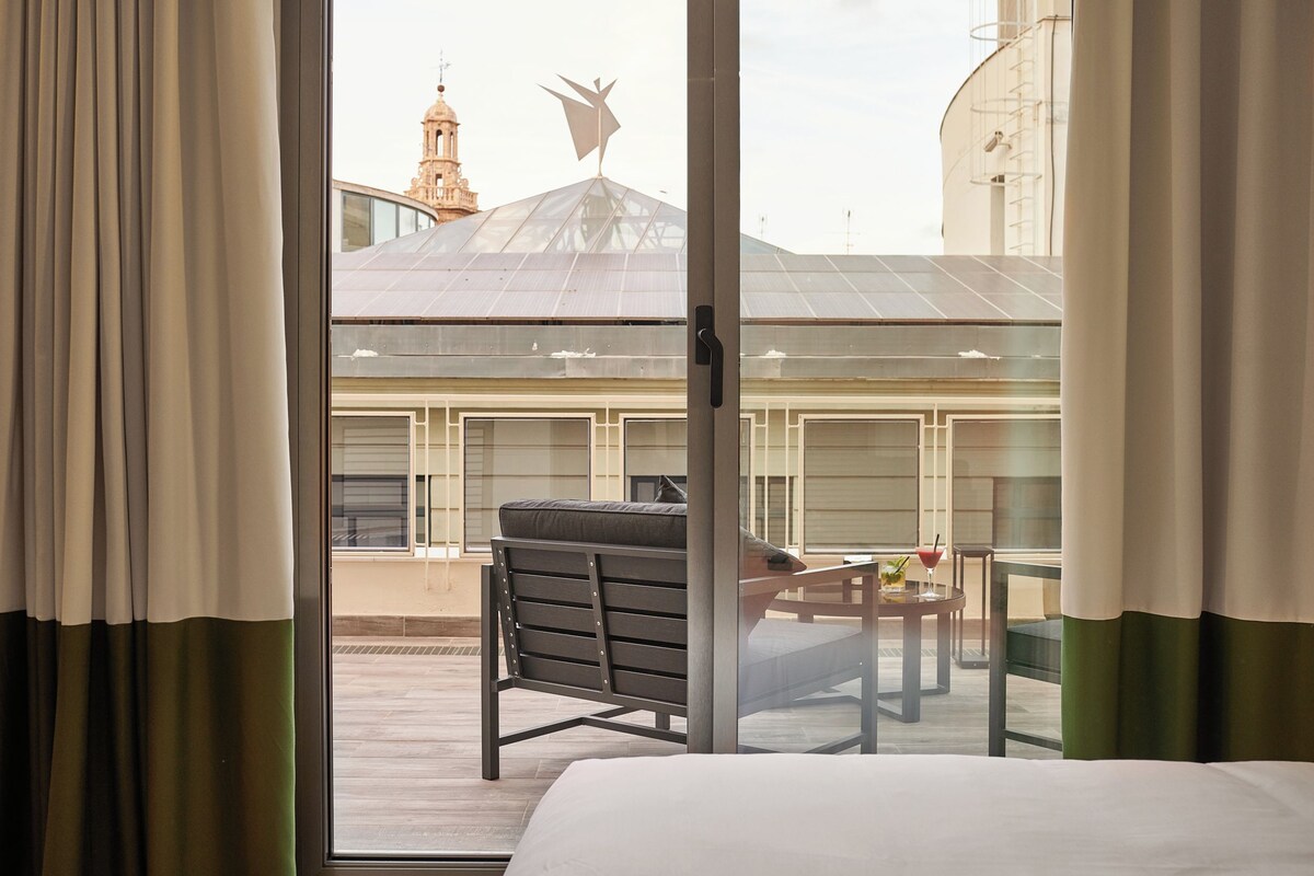 Habitación de hotel con cama, un banco, una mesa con una bebida y vista a un edificio con techo de vidrio.