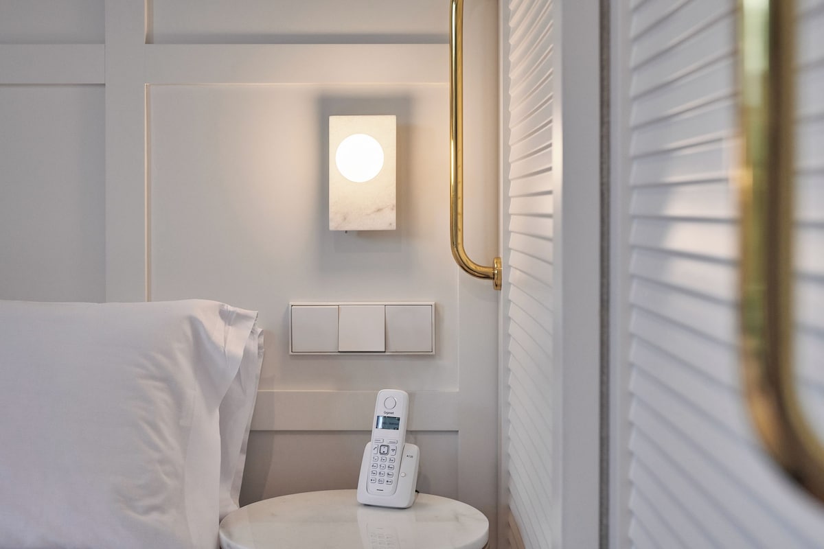 Un baño con inodoro y lavamanos blancos, una luz empotrada en la pared y un cabezal de ducha de mano.
